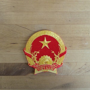 Puede incluir: Un parche bordado en oro con el escudo nacional de Vietnam. El escudo presenta una estrella roja sobre un fondo rojo, rodeada de una corona de espigas de arroz doradas y un engranaje. Las palabras "VIETNAM" están escritas en oro en la parte inferior.