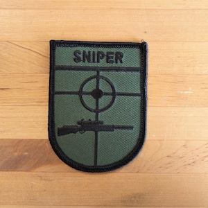 Op de afbeelding: Een groene en zwarte geborduurde patch met het woord "SNIPER" in zwarte letters. De patch heeft een zwart geweer met een richtkijker en een zwart doelwit met een bullseye.