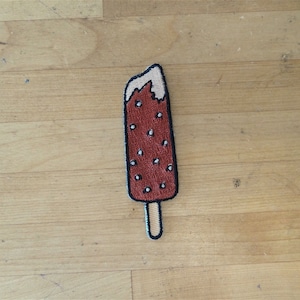 Pode incluir: Um remendo bordado de picolé com um palito, em marrom e branco.