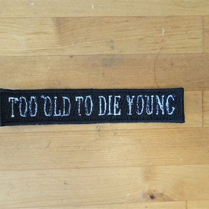 May include: Black embroidered patch with white text that reads "Too Old To Die Young".