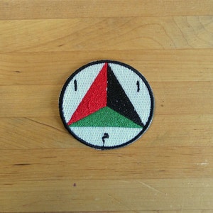 Könnte beinhalten: Ein weißer runder Patch mit einem schwarzen, roten und grünen Dreieck darin. Das Dreieck hat auf jeder Seite eine schwarze Linie und den Text "I" auf jeder Seite. Das Dreieck zeigt nach oben.