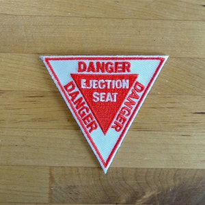 May include: White triangular patch with red embroidered text that reads "DANGER EJECTION SEAT DANGER".