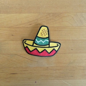 Può includere: Un toppa ricamata di un sombrero giallo e rosso con bordi verdi e bianchi.