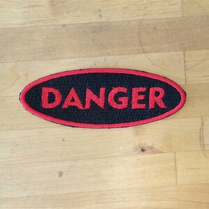 May include: A black oval patch with a red embroidered border and the word "DANGER" in red lettering.