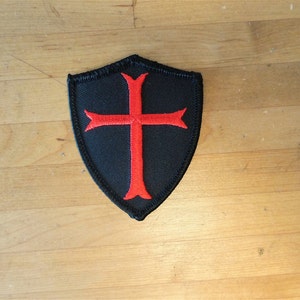 Peut inclure: Un écusson en tissu noir en forme de bouclier avec une croix rouge au centre. La croix a quatre pointes et est un symbole des Templiers.