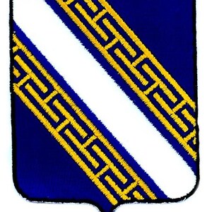 Puede incluir: Parche en forma de escudo con un diseño heráldico. El parche tiene un fondo azul oscuro con una franja diagonal blanca. La franja está bordeada por un patrón geométrico dorado y repetitivo. El parche tiene un borde negro.