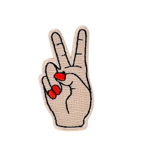 Può includere: Toppa in tessuto beige con una mano ricamata che fa il segno della pace. La mano ha lo smalto rosso.