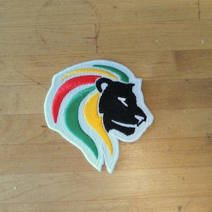 Puede incluir: Un parche blanco con un perfil de cabeza de león negro con una melena roja, amarilla y verde.