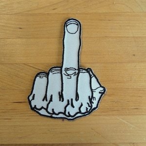 以下が含まれることがあります： 黒い輪郭で刺繍された白い中指のパッチ