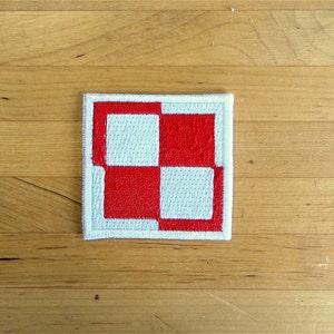 Può includere: Una toppa di tessuto bianco con un motivo a scacchi rosso e bianco. La toppa è quadrata e ha un bordo bianco.