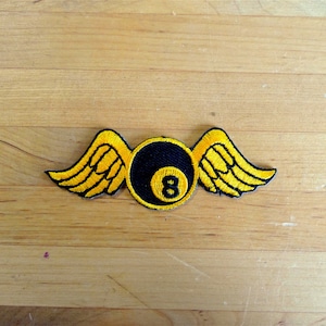 Op de afbeelding: Een geborduurde patch met een zwarte 8-ball met gele vleugels. De 8-ball is zwart met een witte 8 erin. De vleugels zijn geel met zwarte contouren.
