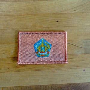 May include: A small, rectangular patch with a peach-colored background and a blue and yellow embroidered emblem. The emblem is a stylized shield with a crown on top.