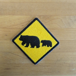 May include: Embroidered patch with a yellow diamond shape and a black border. The patch shows a black silhouette of a bear and a cub, possibly a wildlife warning. Designed for application on clothing or bags, it's a visual alert.