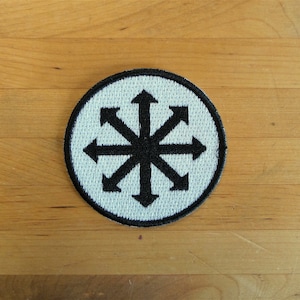 Könnte beinhalten: Ein weißer runder Patch mit einem schwarzen achtzackigen Stern mit Pfeilen, die in alle Richtungen zeigen.