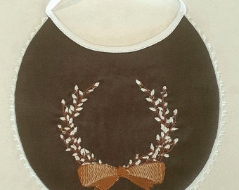 May include: A brown bib with a white lace trim and a brown embroidered wreath with a brown bow.