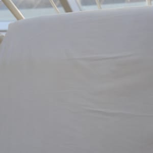 May include: A white sheet draped over a white metal frame, possibly a bed frame. The sheet has subtle wrinkles and folds, suggesting it is made of a soft fabric. The frame is visible in the corner, with a window in the background.