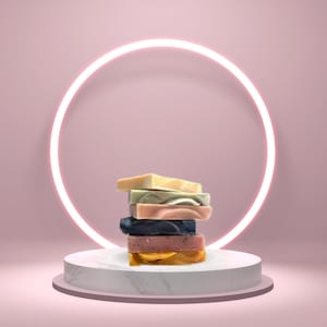 May include: A stack of five different colored handmade soap bars on a white marble pedestal with a glowing neon circle behind it.