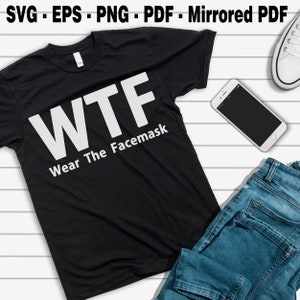 May include: Black t-shirt with the white text "WTF Wear The Facemask." The shirt is displayed with blue jeans, a smartphone, and a white sneaker on a white wooden surface. The top of the image has the text "SVG - EPS - PNG - PDF - Mirrored PDF."