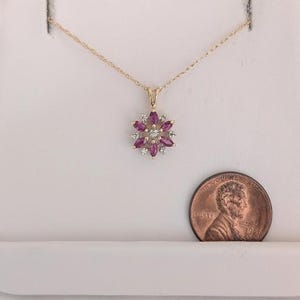 Könnte beinhalten: Eine goldfarbene Halskette mit einem blütenförmigen Anhänger. Der Anhänger ist mit rosa Edelsteinen und kleinen, klaren Steinen besetzt. Eine Münze liegt neben der Halskette zum Größenvergleich.