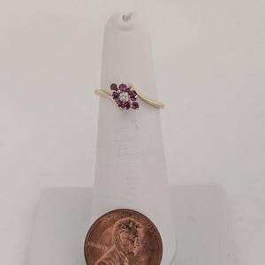 May include: A gold ring featuring a floral design with a central diamond and surrounding red gemstones. The ring is displayed on a white stand, with a penny in the foreground for size reference. The ring's band has a delicate, swirling design.