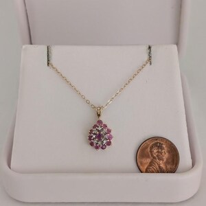 May include: A gold-colored necklace with a pendant featuring a cluster of red gemstones and smaller white gemstones. The necklace is displayed in a white jewelry box, with a penny for size comparison.