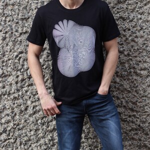 May include: A black t-shirt with a white graphic of a circular pattern. The person wearing the shirt is standing in front of a textured wall.