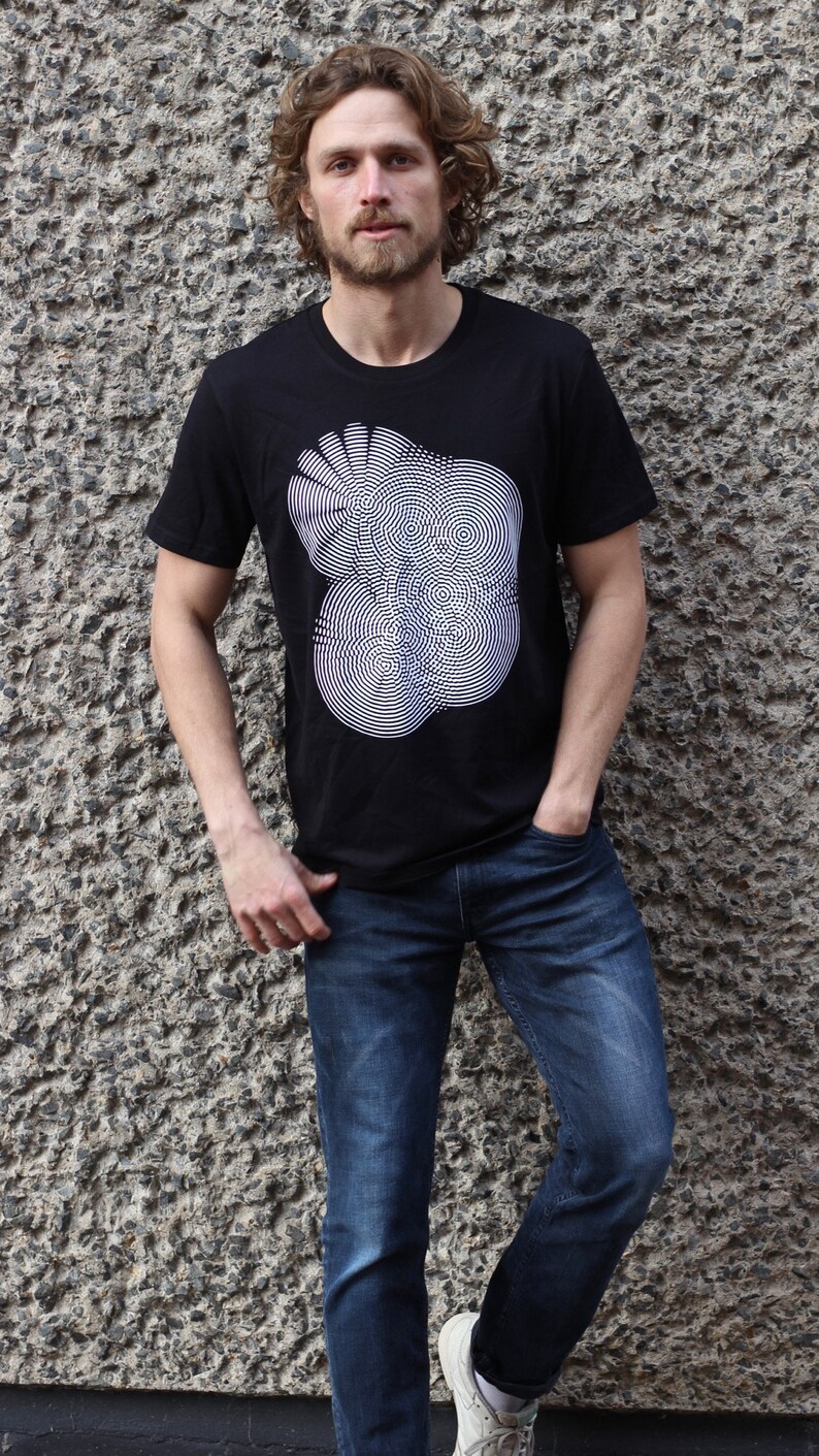 May include: A black t-shirt with a white graphic of a circular pattern. The person wearing the shirt is standing in front of a textured wall.