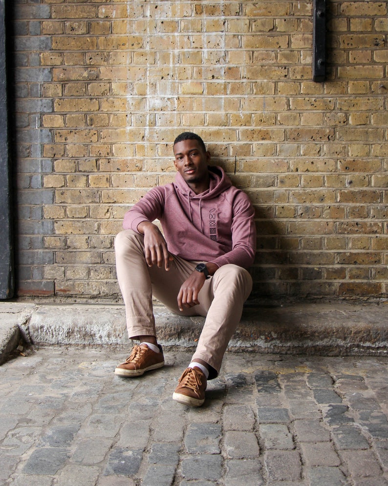 Op de afbeelding: Een man in een roze hoodie met een geometrisch ontwerp zit met gekruiste benen op een geplaveide weg. Hij draagt ​​een kaki broek en bruine schoenen.