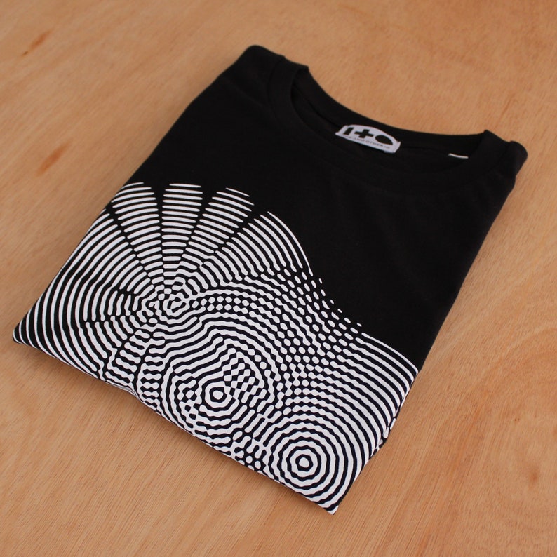 May include: Black t-shirt with a white graphic print of a circular pattern. The pattern is made up of concentric circles and lines.