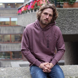 Op de afbeelding: Een man draagt een bordeauxrode hoodie met een geometrisch ontwerp op de voorkant. Hij zit op een betonnen rand met zijn handen in zijn schoot.