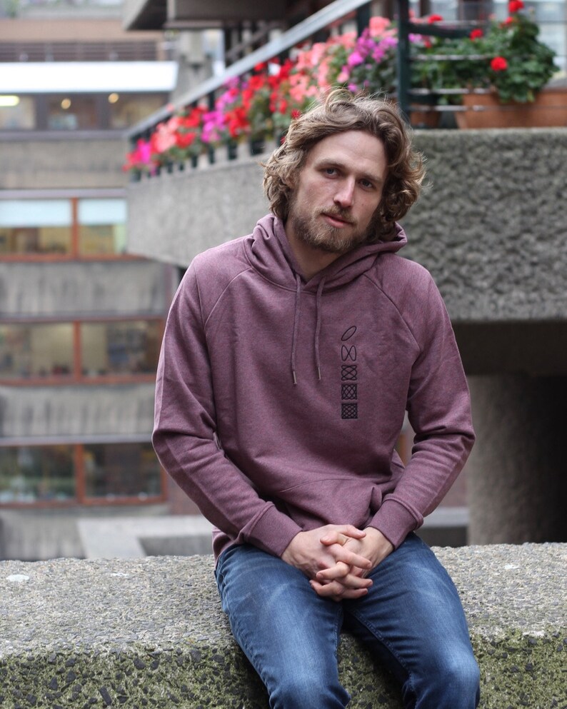 Op de afbeelding: Een man draagt een bordeauxrode hoodie met een geometrisch ontwerp op de voorkant. Hij zit op een betonnen rand met zijn handen in zijn schoot.