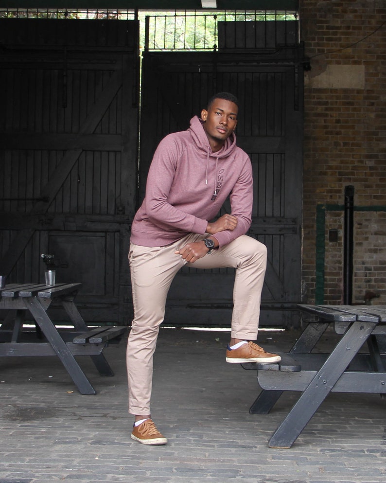 Op de afbeelding: Een man draagt een mauve hoodie met het woord "OMSG" erop gedrukt, een kaki broek en bruine schoenen. Hij staat voor een zwarte metalen poort en een bakstenen muur.