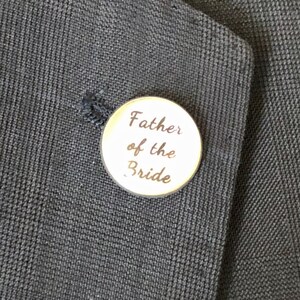 May include: A round, gold-colored pin with the text "Father of the Bride" in a script font. The pin is attached to a dark navy blue suit jacket. The jacket has a subtle textured pattern.