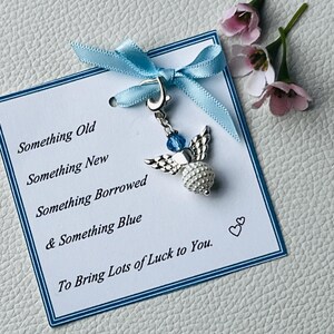 Op de afbeelding: Een bruiloft-thema bedel met een zilveren engel hanger, blauwe kraal accenten en een lichtblauw lint. De bedel is bevestigd aan een kaart met de tekst "Something Old, New, Borrowed, & Blue. To Bring Lots of Luck to You."