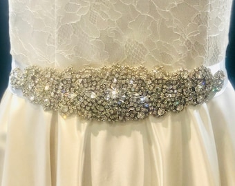 May include: A white satin wedding dress with a sparkling rhinestone belt. The belt is a wide band of clear rhinestones in various shapes and sizes, set on a white satin ribbon. The bodice is white lace.