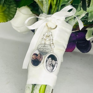 Puede incluir: Un encanto de ramo de novia de satén blanco con un encanto de corazón plateado que dice "un pedazo de mi corazón está en el cielo". El encanto está unido a una cadena plateada con cuentas de perlas. Dos encantos de fotos de forma ovalada están unidos a la cadena, cada uno contiene una foto en blanco y negro de una pareja.