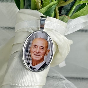 May include: A silver oval pendant featuring a photograph of a man in a suit. The pendant is attached to a white ribbon.