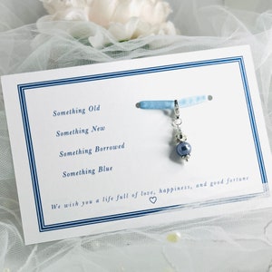 Puede incluir: Una tarjeta blanca con borde azul y el texto "Something Old, Something New, Something Borrowed, Something Blue." Una cinta azul con un dije plateado con una perla azul oscuro está unida a la tarjeta. El texto "We wish you a life full of love, happiness, and good fortune" está impreso debajo de la lista. Un símbolo de corazón está impreso debajo del texto.