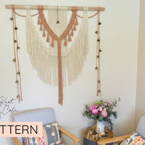 May include: A macrame wall hanging with a peach and white color scheme, featuring a geometric pattern and tassels. The hanging is adorned with a string of small bells.