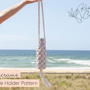 Pode incluir: Um porta-garrafas de macramé branco com uma borla pendurada na parte inferior. O porta-garrafas está sendo segurado por uma mão contra um fundo de praia e oceano. O texto "Macrame Bottle Holder Pattern" está na parte inferior da imagem.