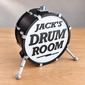 May include: A black and white decorative drum-shaped sign featuring the text "JACK'S DRUM ROOM" in raised black lettering on a white circular face. The object has silver-coloured hardware accents and is supported by two silver-coloured legs.