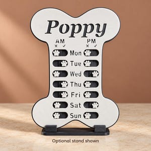 Pode incluir: Um rastreador de alimentação para animais de estimação em forma de osso com superfície branca e borda preta. Apresenta o nome Poppy no topo, com colunas AM e PM para cada dia da semana de Mon a Sun. Deslizadores em forma de pata marcam o uso.