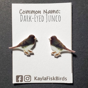 May include: A pair of earrings featuring a dark-eyed junco bird design. The birds are depicted in shades of brown, white, and green. The earrings are made of polymer clay and are approximately 1 inch in size. The card reads "Common Name: DARK-EYED JUNCO" and includes social media handles for KaylaFiskBirds.