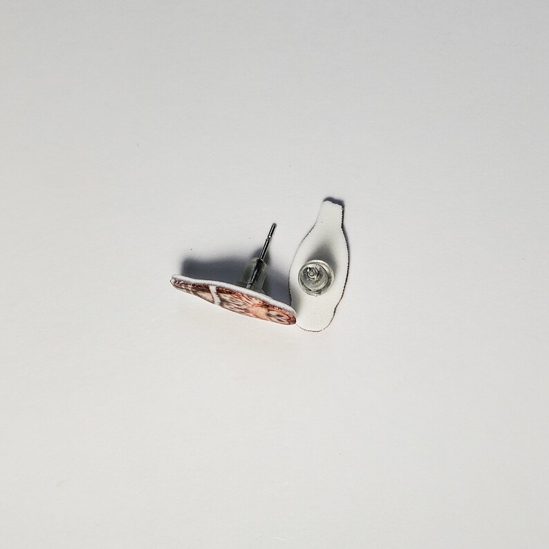 K&ouml;nnte beinhalten: Ein Paar Ohrstecker mit einem braunen und wei&szlig;en Design. Die Ohrringe sind aus Metall und haben einen Stiftverschluss.