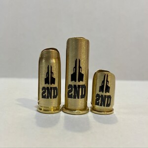 May include: Three gold-colored metal bullet casings with black printed designs. The casings are different sizes and each has a silhouette of two pistols and the word "2ND" printed on them.