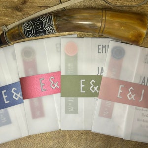 Può includere: Cinque buste bianche con un nastro decorativo e le iniziali "E & J" stampate su ciascuna. Le buste sono disposte in fila su una superficie di legno. La busta superiore ha il testo "Emily & James" stampato su di essa.