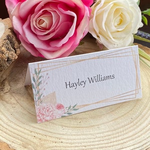 May include: A folded place card with the name "Hayley Williams" printed in black. The card has a gold geometric border and floral watercolor accents in pink and green. The card is displayed on a wooden slice with pink and white roses.