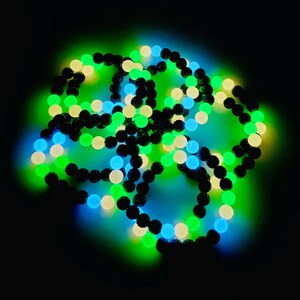 May include: A collection of glowing bead necklaces in a dark setting. The beads are a mix of black, green, blue, and off-white, creating a vibrant, luminous effect. The necklaces are arranged in a tangled, circular pattern.