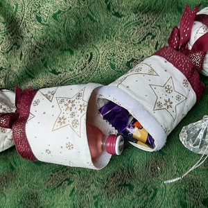 May include: A white fabric Christmas cracker with gold stars and a burgundy ribbon. The cracker is filled with small gifts, including a bottle of perfume, chocolate, and a small toy.