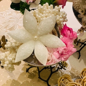 May include: A white, sparkly, star-shaped flower sits on a silver stand with a black branch design. The flower is surrounded by pink flowers, white pearl beads, and white flowers. The stand is on a white and gold patterned surface.